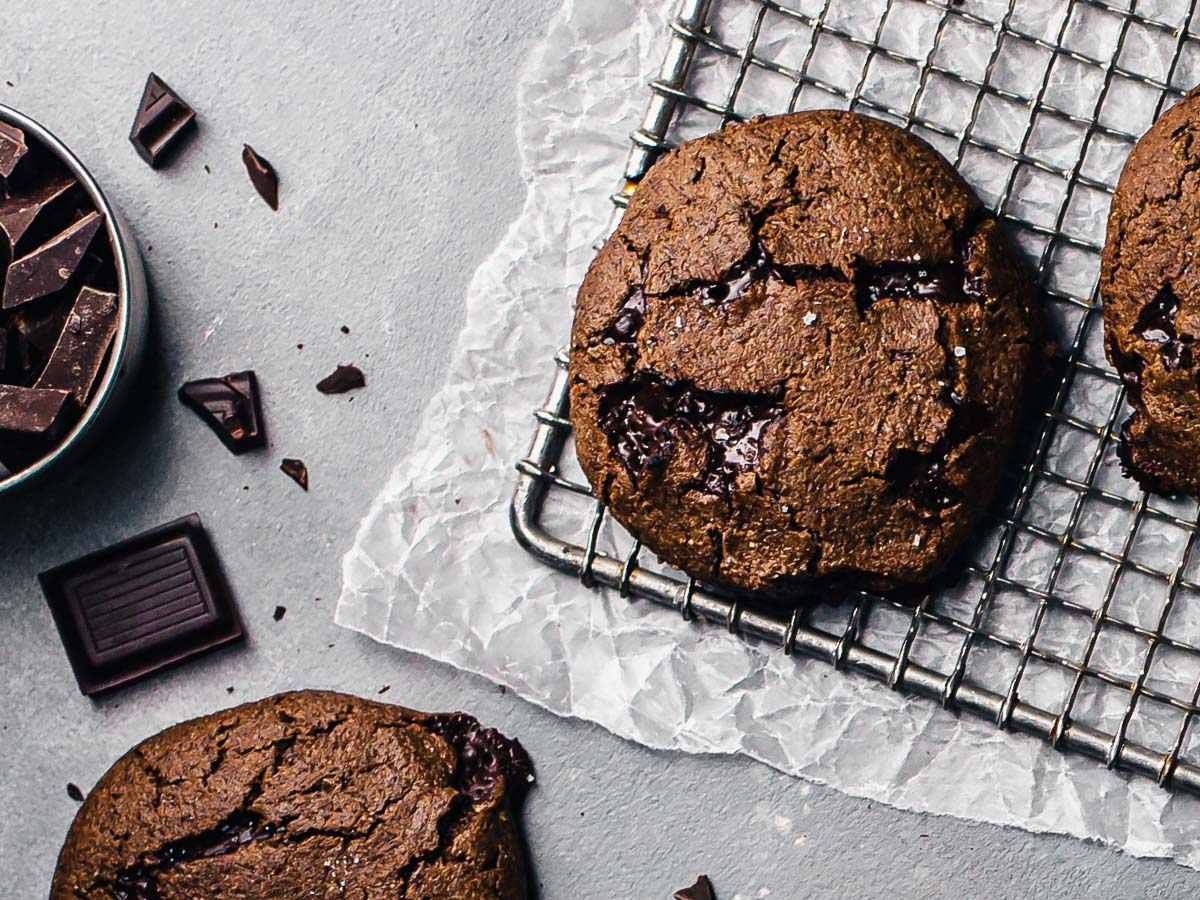 One vegan double chocolate espresso cookie on a cooling rack on a concrete counter, with chocolate chunks and more cookies surrounding.