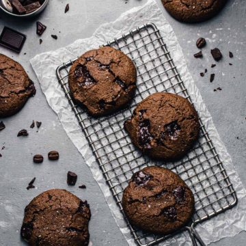 Three vegan double chocolate espresso cookies on a cooling rack on a concrete counter, with chocolate chunks and more cookies surrounding.