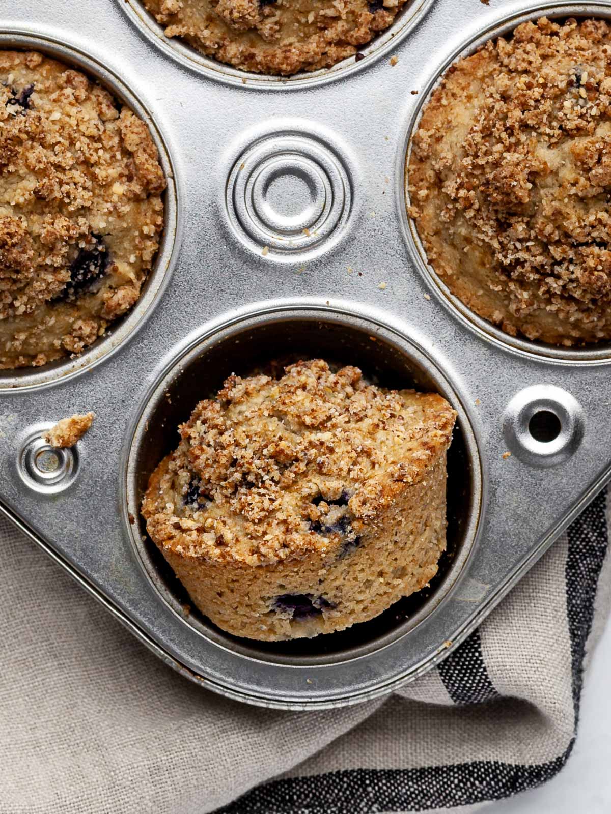 One vegan low-fodmap blueberry muffin on its side in a muffin tin with two muffin tops framing..