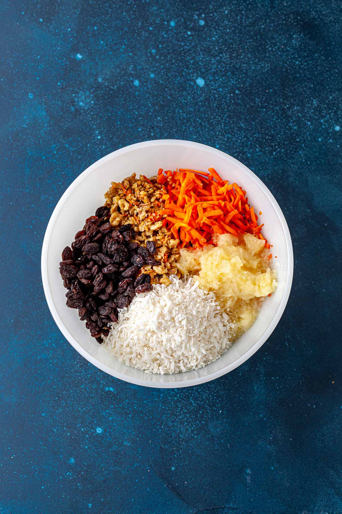 Grated carrots, chopped walnuts, raisins, shredded coconut, and crushed pineapple in a bowl.