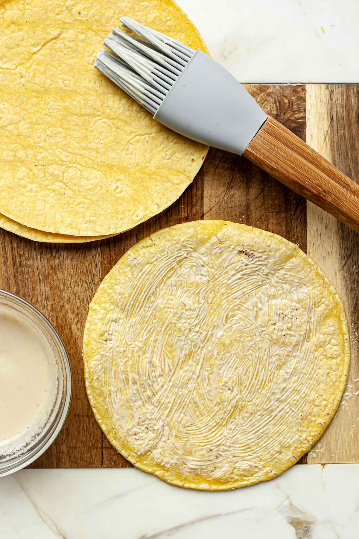 Tahini mixture being brushed on the other side of a tortilla with a stack of tortillas in the background.