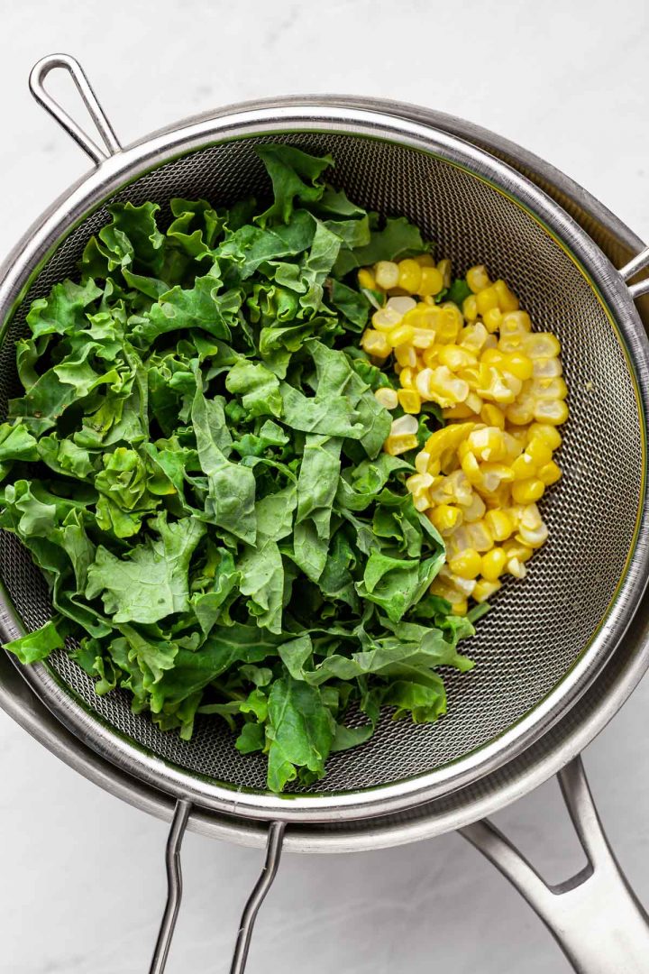 Overhead view of uncooked kale and corn in a mesh strainer on top of a pot of beans.