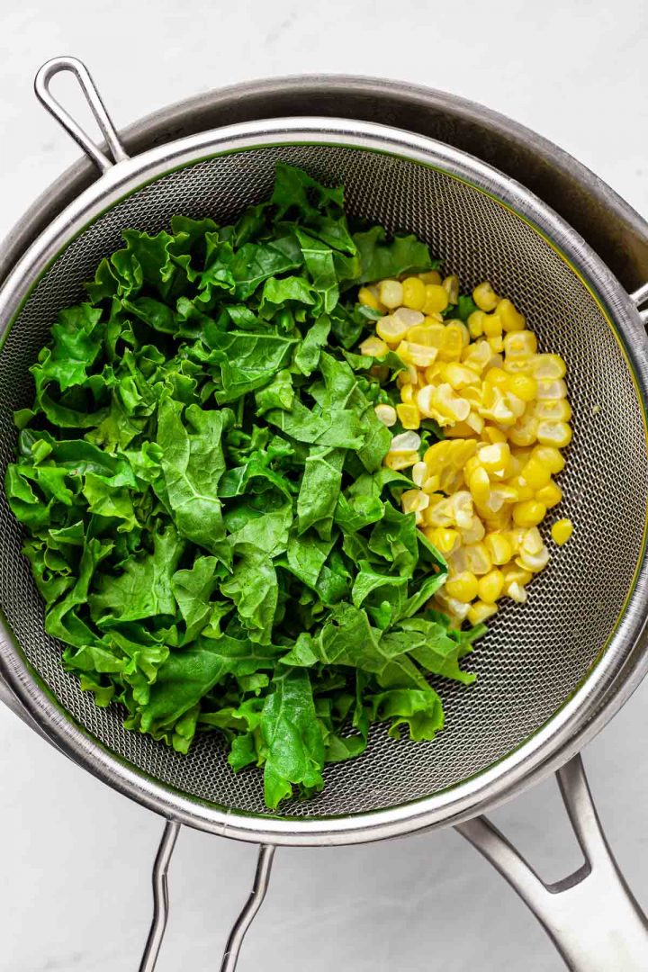 Overhead view of cooked kale and corn in a mesh strainer on top of a pot of beans.