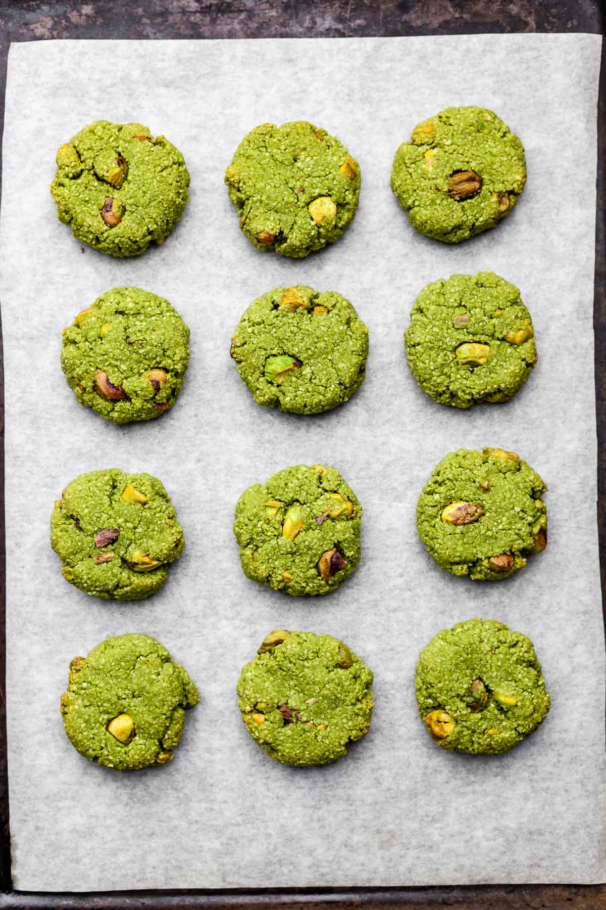 Overhead view of a parchment-lined cookie sheet with twelve evenly-spaced vegan matcha cookies after being baked.