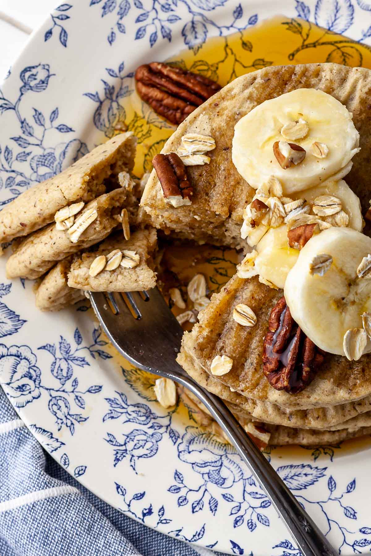 Overhead view of a stack of vegan oat flour pancakes topped with banana slices, toasted oats, and chopped pecans, drizzled with pure maple syrup. To the left, there is a fork holding three pieces of pancakes. The meal is served on a blue and white floral plate.