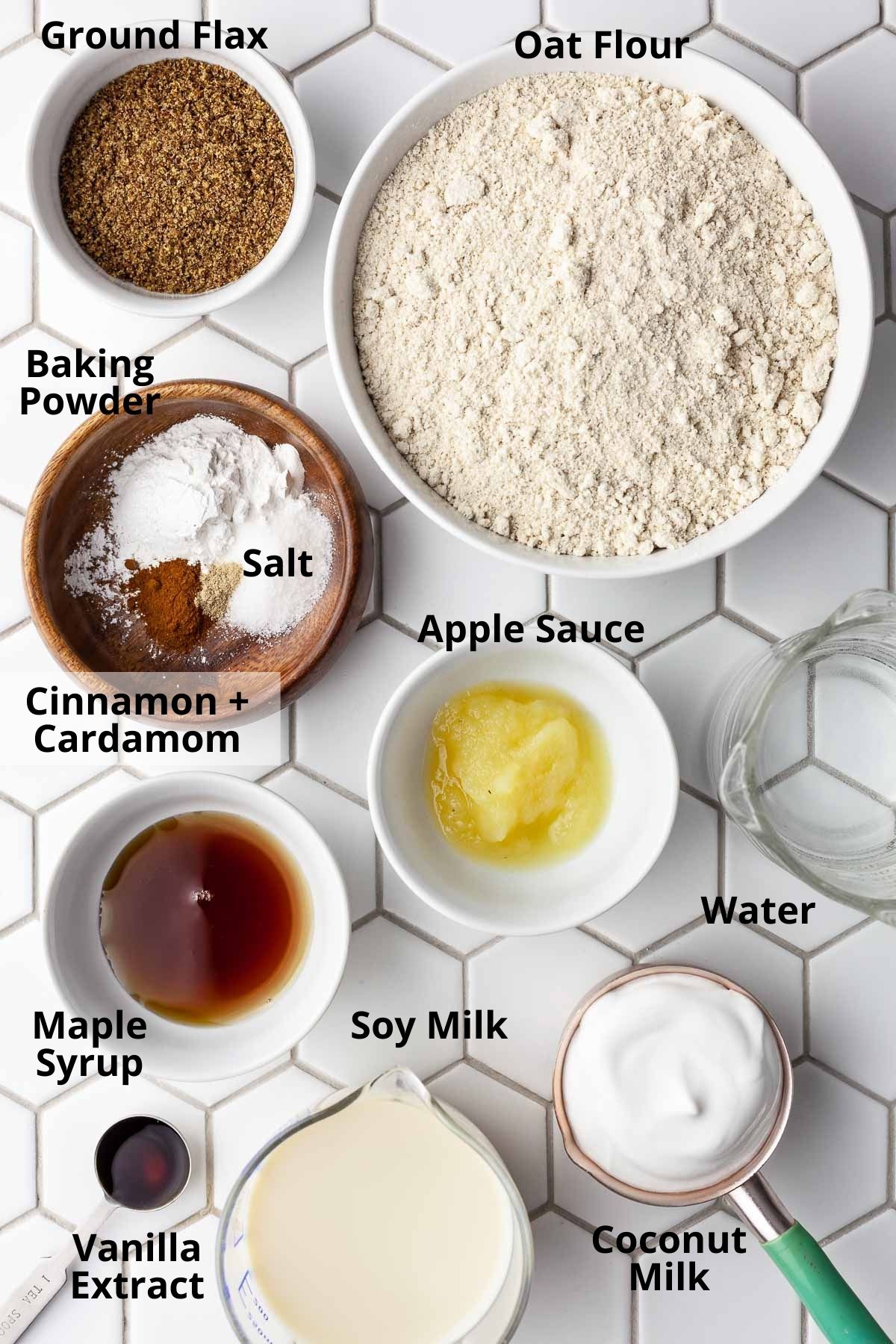 Overhead view of individual oil-free vegan oat flour pancake ingredients labeled and separated into bowls on a white hexagon tile backdrop.
