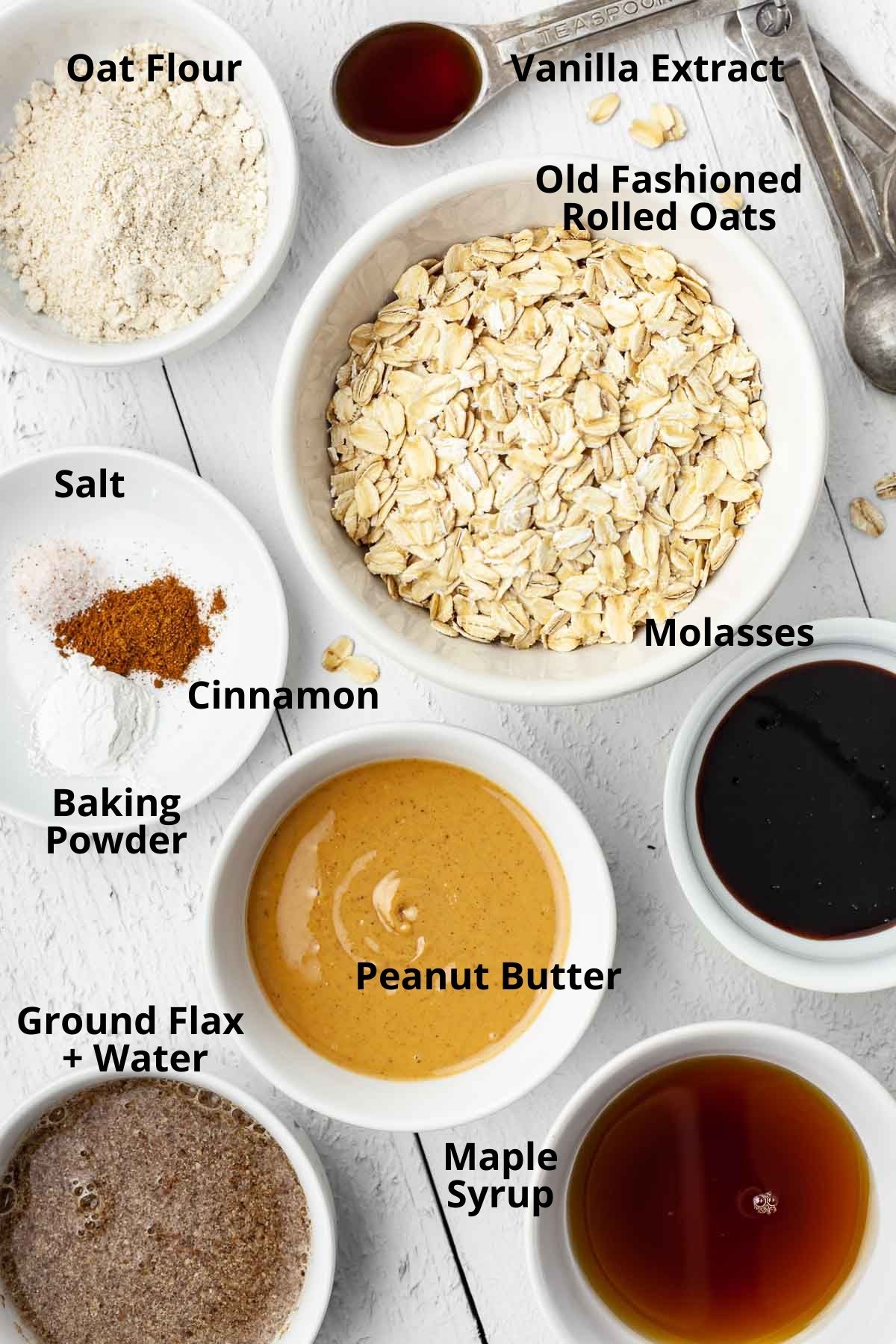 Overhead view of individual vegan peanut butter oatmeal cookie ingredients labeled and separated into bowls on a white wood plank table.