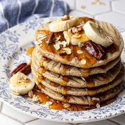 Stack of five vegan oat flour pancakes topped with banana slices, toasted oats, and chopped pecans, drizzled with pure maple syrup, and served on a blue and white floral plate.