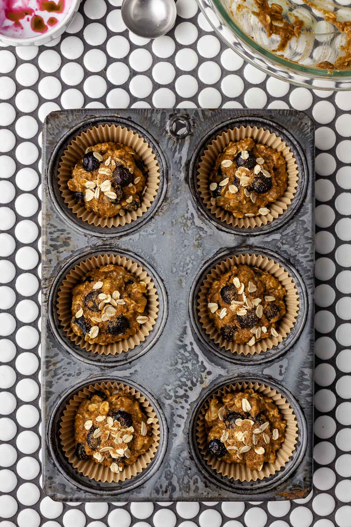 Muffin tin holding banana blueberry oatmeal muffin batter with added blueberries, rolled oats, and coconut sugar on top.